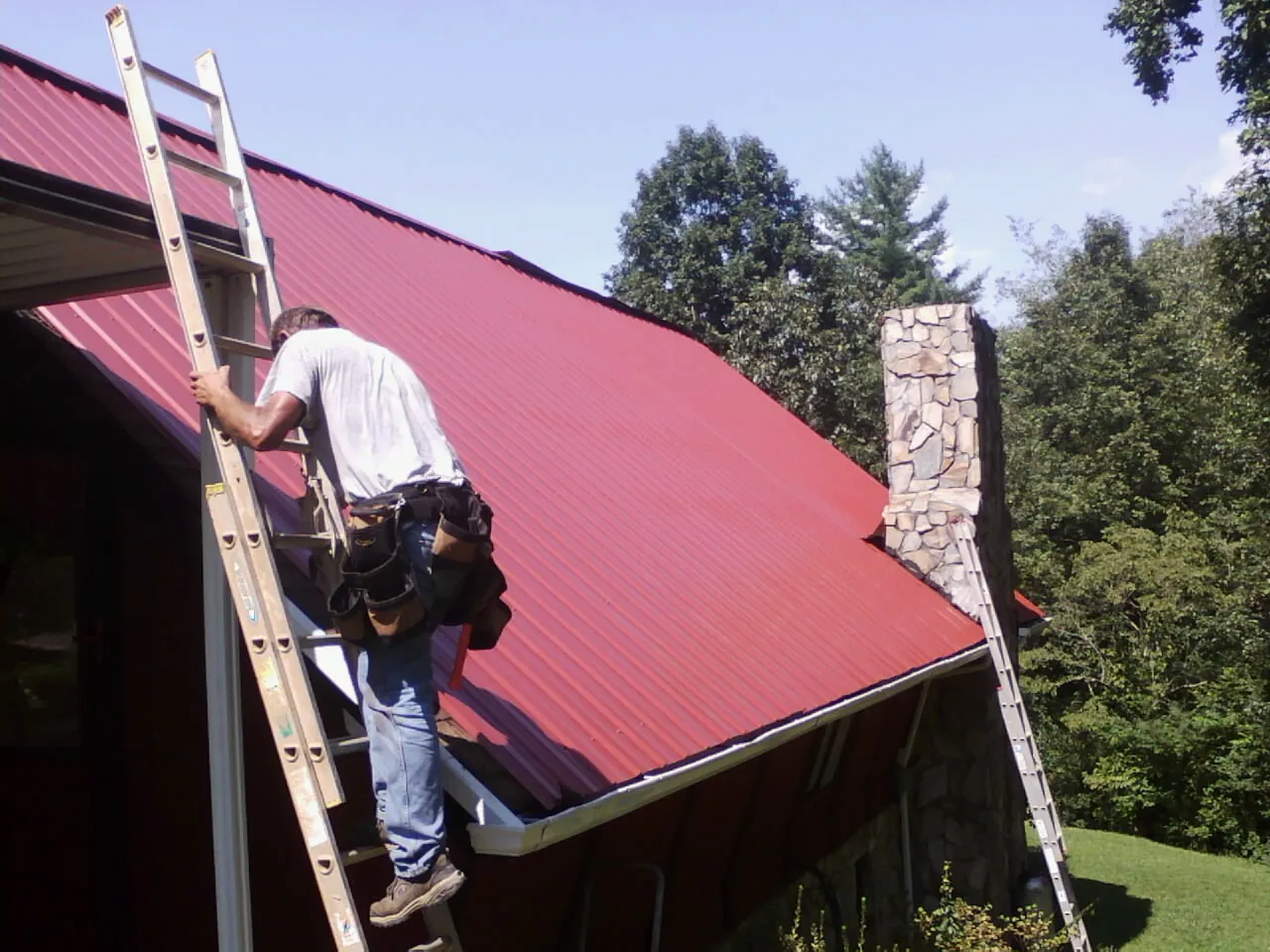 Roofer on a ladder beside a red metal roof and stone chimney — Soffit & Fascia Repair by Roofing Brothers in Lower Salem
